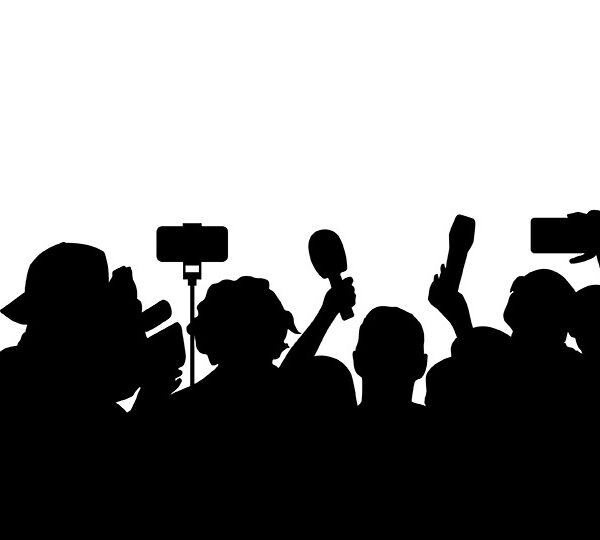 Bildbeschreibung: Das Bild zeigt die Silhouetten einer Gruppe von Medienvertretern, darunter Fotografen und Reporter, die Kameras und Mikrofone halten, vermutlich bei einer Pressekonferenz oder einem ähnlichen Ereignis.
