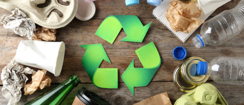 Das Bild zeigt verschiedene Arten von Müll, die um ein großes grünes Recycling-Symbol auf einer hölzernen Oberfläche verstreut sind.