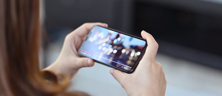 Bildbeschreibung: Eine Person hält ein Smartphone quer in den Händen und schaut ein Video mit unscharfem Stadthintergrund.